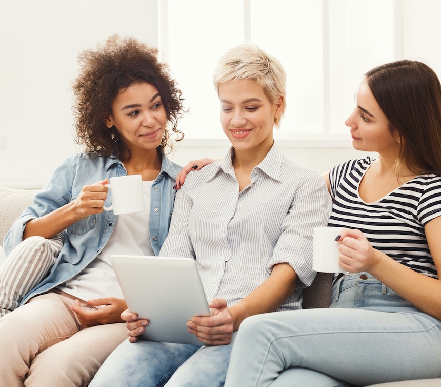 Three happy female friends using tablet and drinking coffee