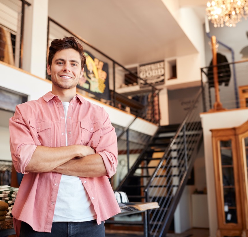 Portrait Of Smiling Male Owner Of Fashion Store Standing In Front Of Clothing Display