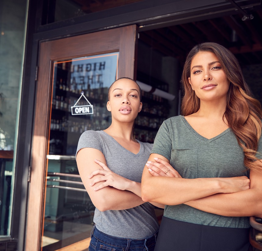 Portrait Of Two Women Starting New Coffee Shop Or Restaurant Business Standing In Doorway