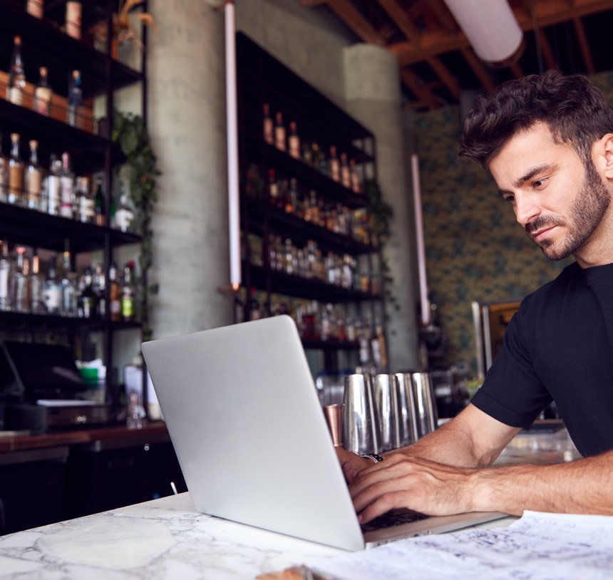Male Owner Of Restaurant Bar Sitting At Counter Working On Laptop